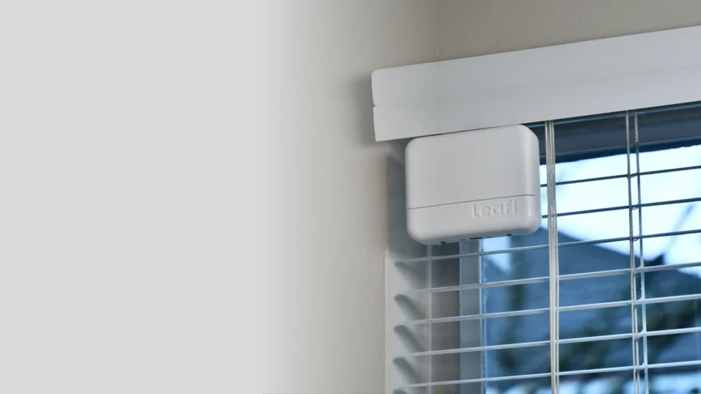 A white smart device mounted above window blinds, designed for automated control, with "Leafi" logo visible on the front.