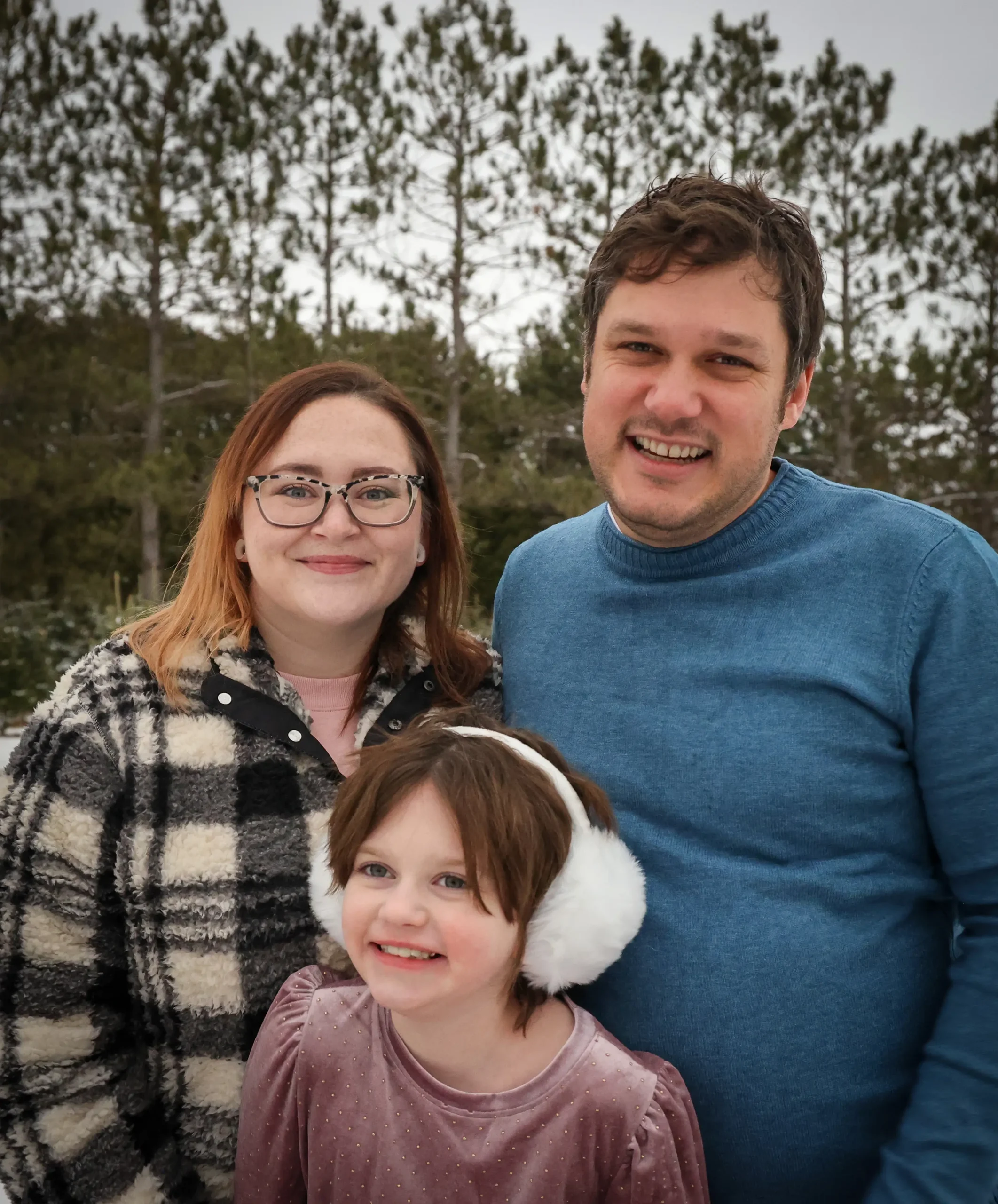 A family, dressed warmly for winter, stands together outdoors with tall evergreen trees in the background.