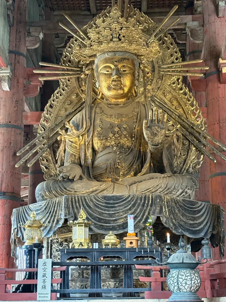 Large, golden statue of a seated bodhisattva, adorned with elaborate headpiece and ornate details, displayed on a platform with offerings and sacred objects in a temple.