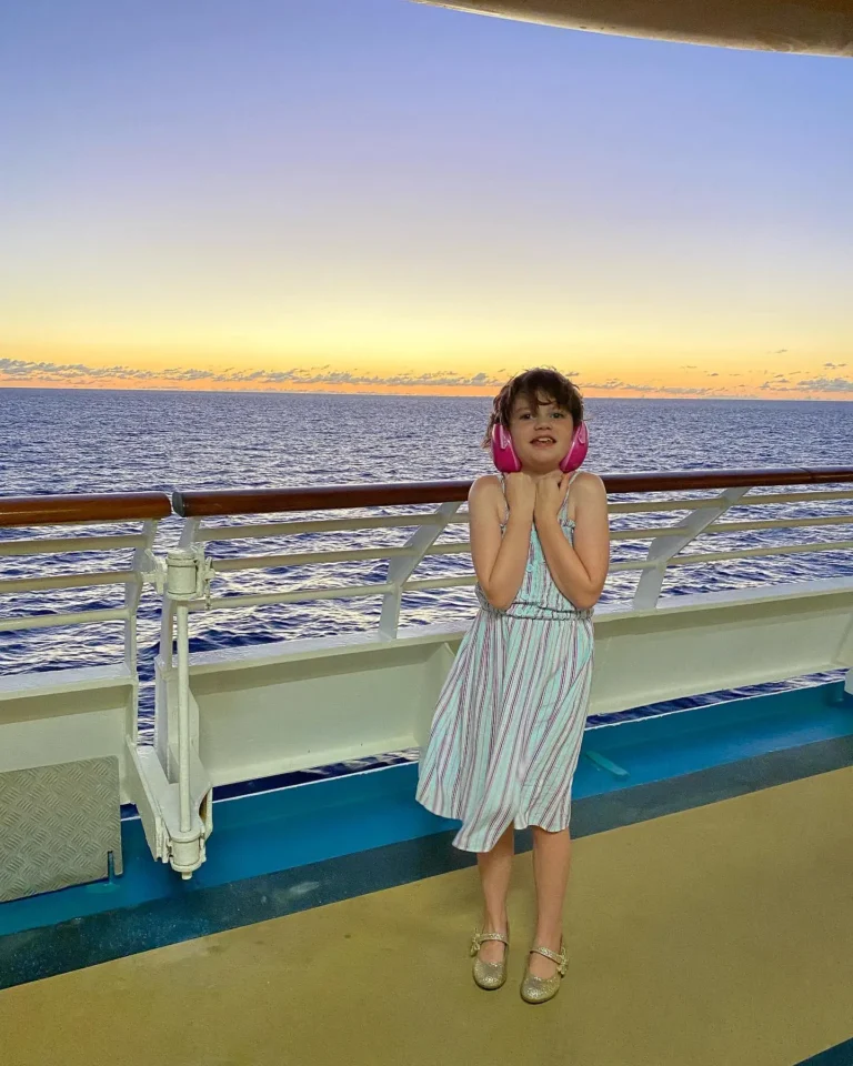 A child stands on a cruise ship deck, wearing headphones and a striped sundress, with a stunning sunset over the ocean behind her.
