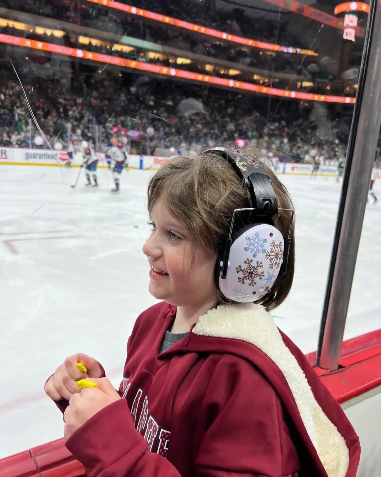 A child with ear protection watches a hockey game, wearing a maroon hoodie, while holding a bright yellow object.