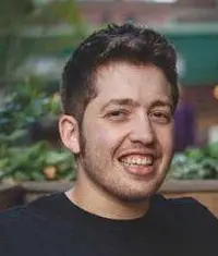A person with dark, styled hair is sitting outdoors, surrounded by greenery, wearing a black shirt and appearing relaxed.