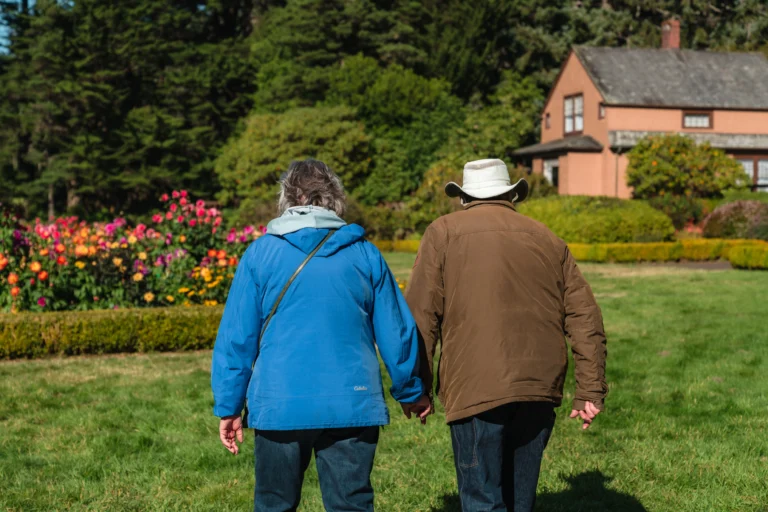A couple walks hand in hand through a garden, surrounded by colorful blooming flowers and a historic house in the background.