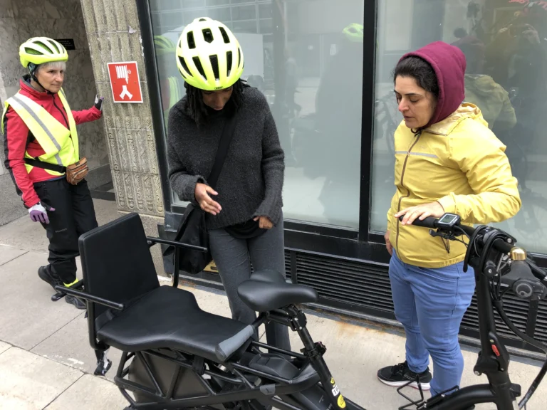 Three individuals in bright helmets engage in a discussion near a bike, with a bicycle seat visible and a pedestrian sign in the background.