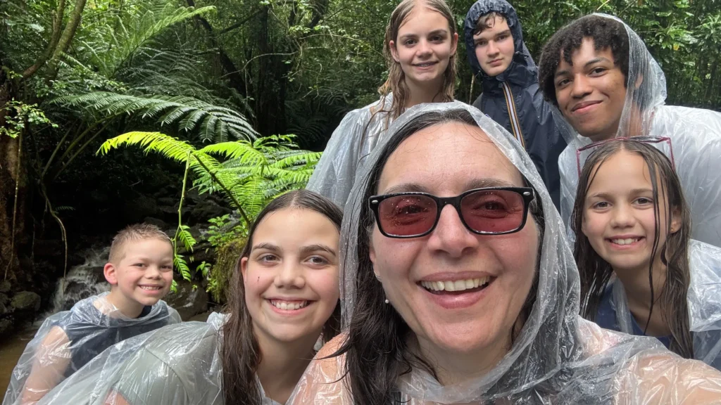 A group of eight people wearing clear ponchos stands in a lush forest setting, with ferns and a creek visible in the background.
