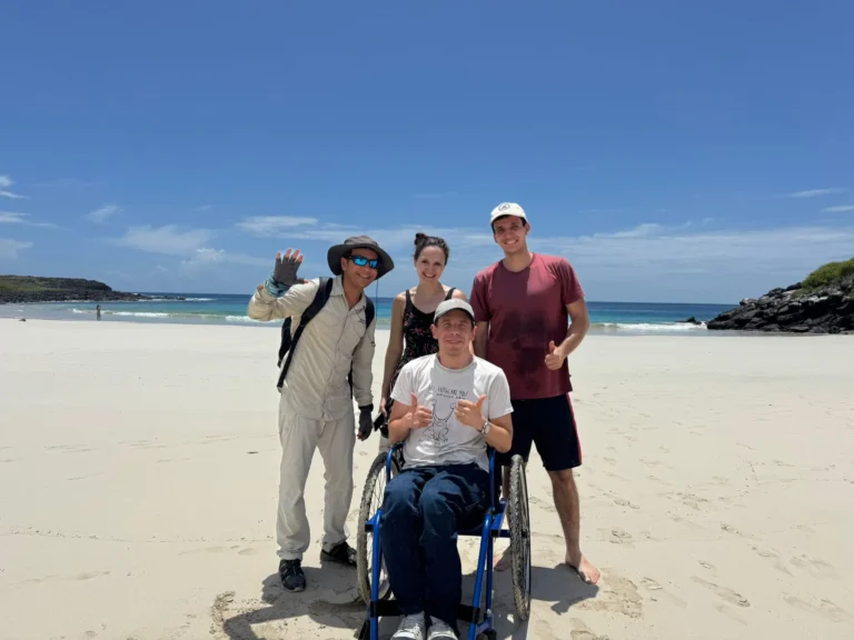 Four people pose and smile on a sunny beach. One man is in a wheelchair, while the others stand around him. The ocean and blue sky are in the background, with some green hills at the edges.