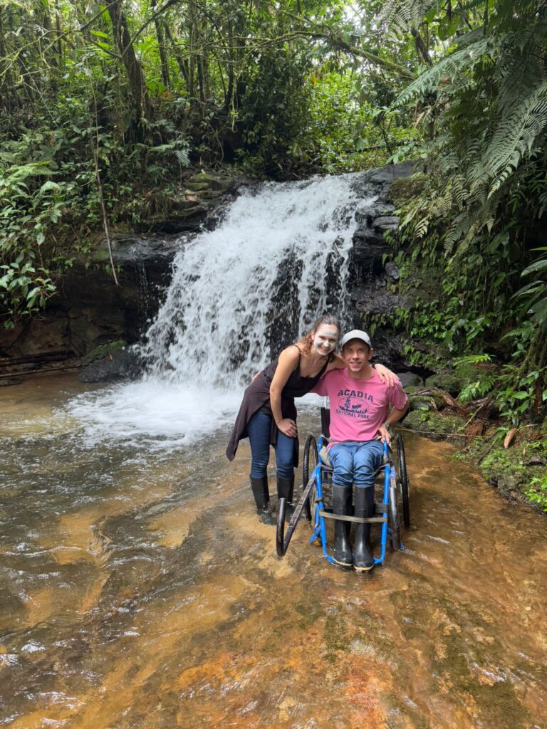 A woman and a man in a wheelchair smile together in front of a small waterfall surrounded by lush green foliage. They are standing in shallow water and both wear rubber boots.