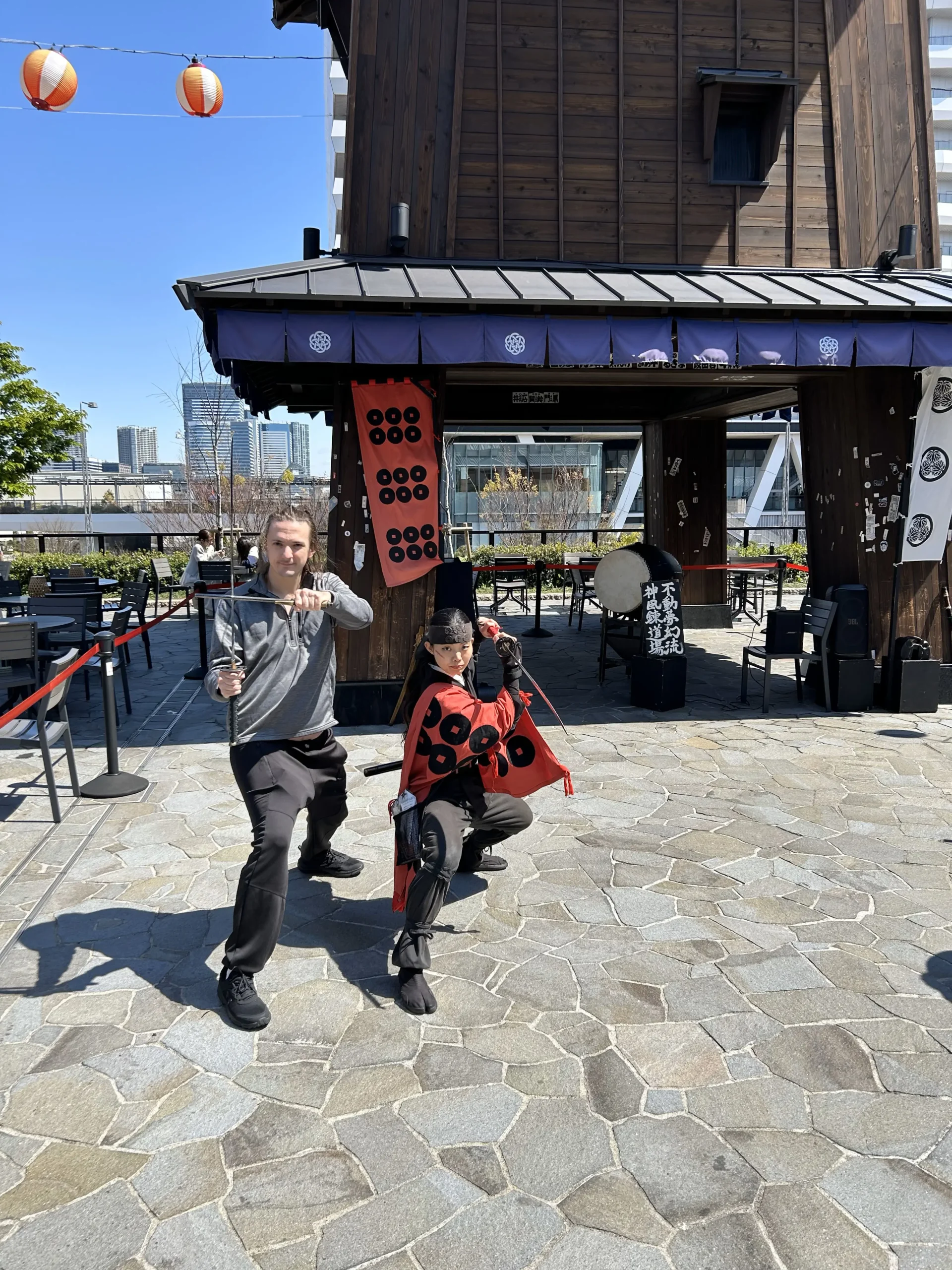 Two people dressed in traditional Japanese warrior attire, holding swords, pose on a stone-paved plaza outside a wooden building.