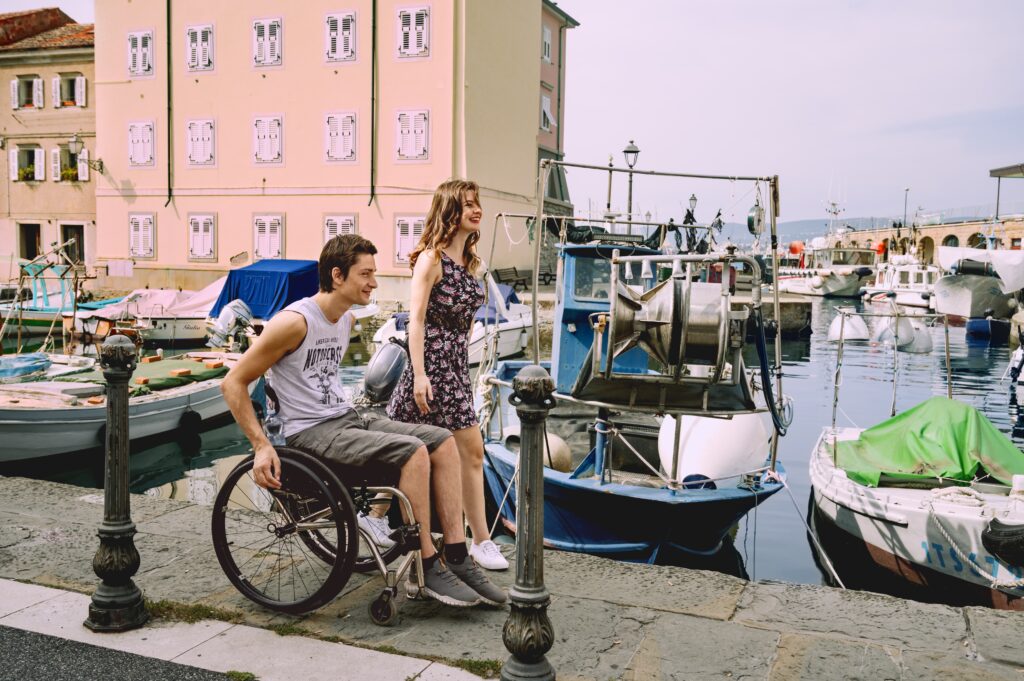 A smiling man in a wheelchair and a woman in a floral dress stroll by a marina. Colorful boats and pastel buildings create a cheerful, relaxed vibe.