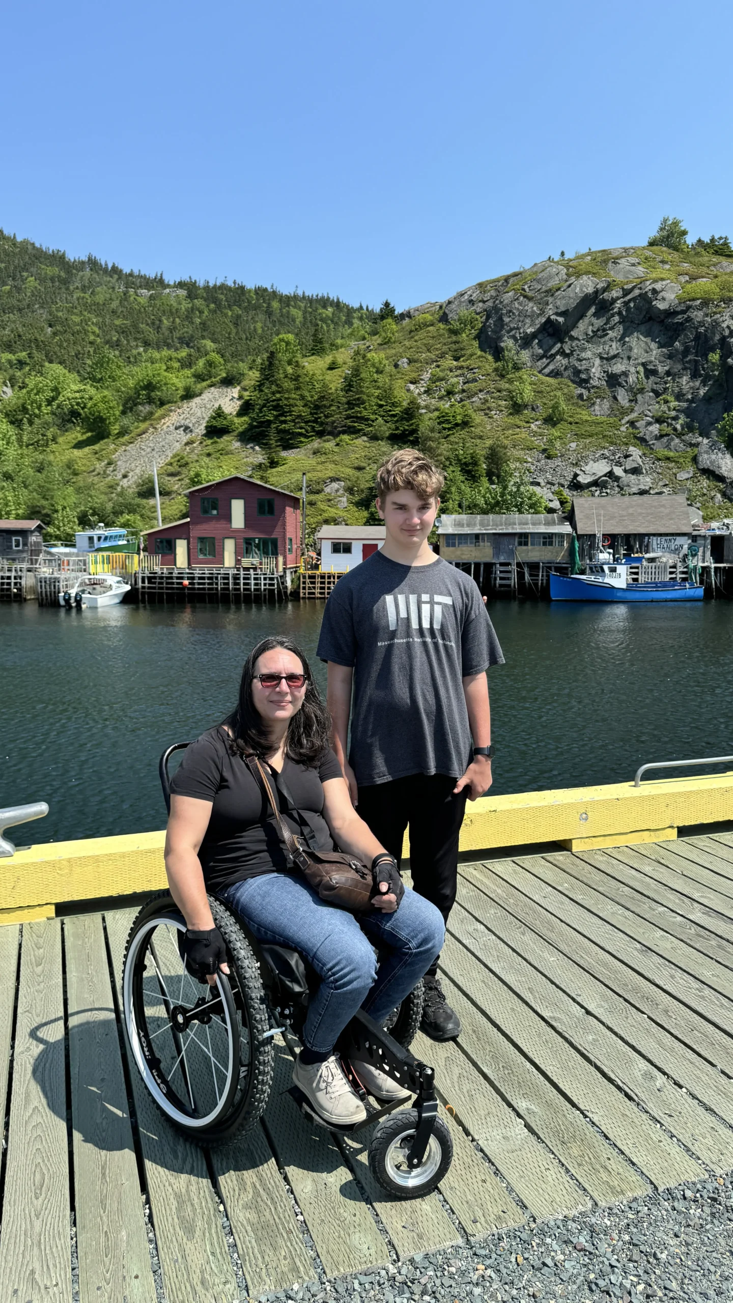 A woman in a wheelchair and a standing boy pose together on a sunny day by the water, with colorful buildings and green hills in the background.