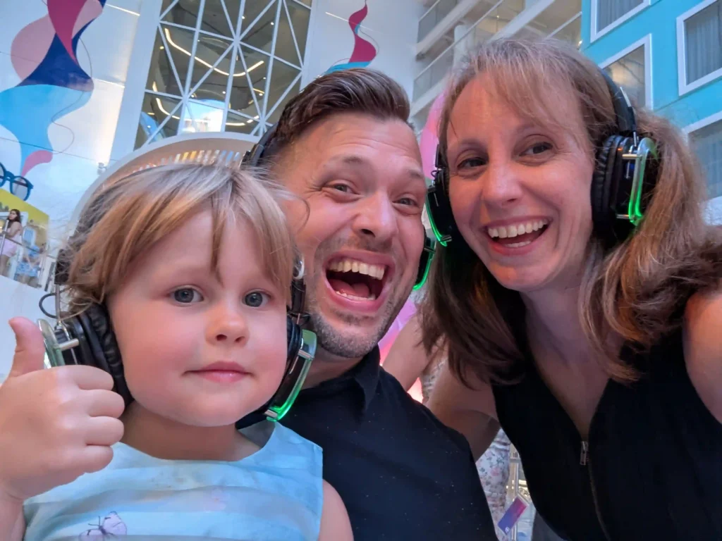 A smiling man and woman, and a young girl giving a thumbs up, all wearing headphones at an indoor event with colorful decorations and tall windows in the background.