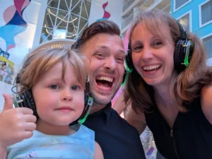 A smiling man and woman, and a young girl giving a thumbs up, all wearing headphones at an indoor event with colorful decorations and tall windows in the background.
