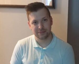 A person with short brown hair and a trimmed beard is wearing a light blue polo shirt, sitting indoors against a beige wall with a framed picture in the background.