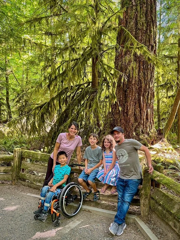 A group of adults and children relax by a large tree in a lush, green forest, enjoying a sunny day outdoors.