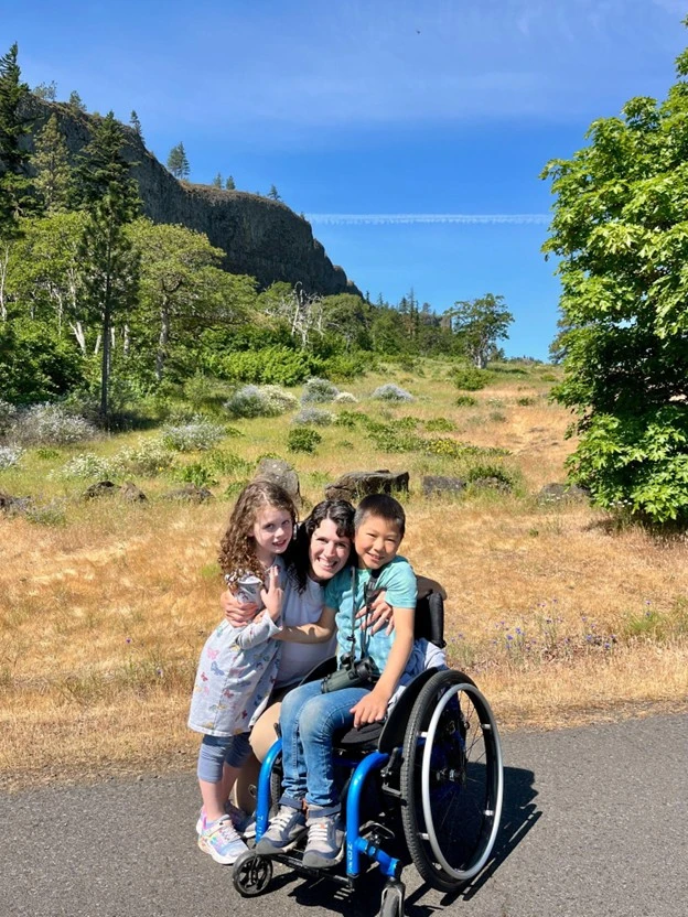 Three children embrace joyfully outdoors, with lush greenery and a rocky hillside in the background under a clear blue sky.