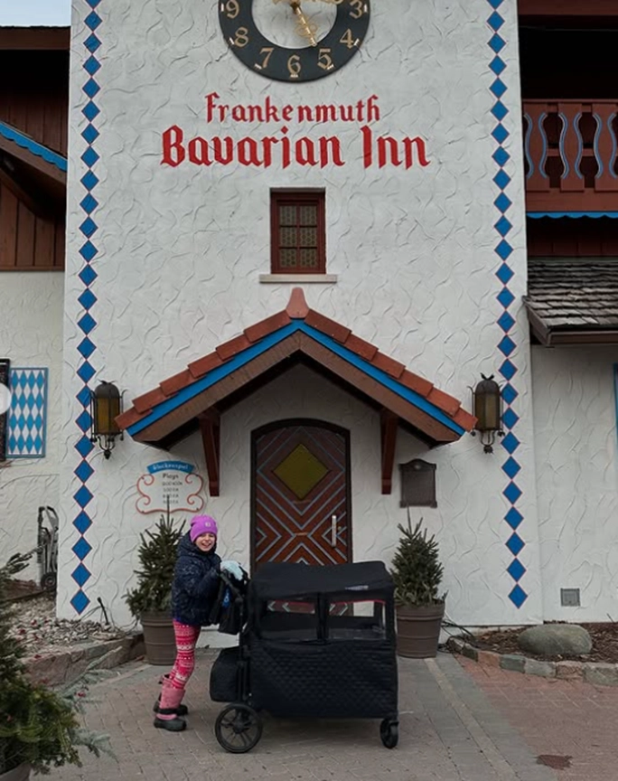A child in colorful leggings pushes a black stroller in front of the Bavarian Inn, featuring a decorative clock and vibrant architecture.