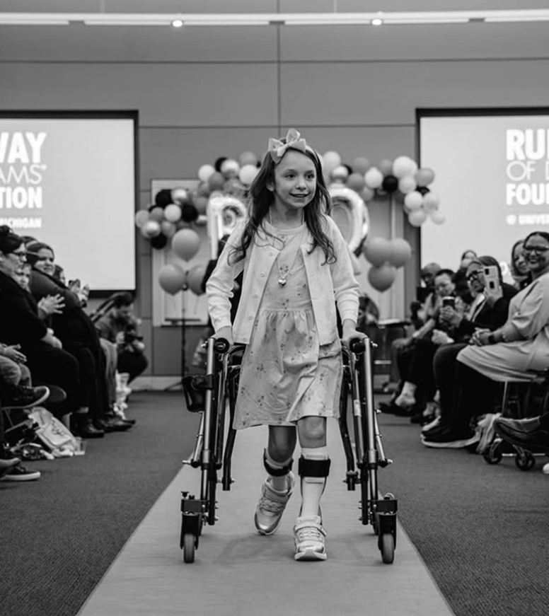A young girl confidently walks down a runway using mobility aids, surrounded by an audience and colorful balloons.