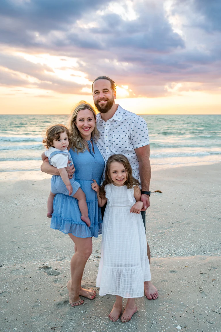 A family of four stands on a beach at sunset, with gentle waves in the background and colorful clouds above.