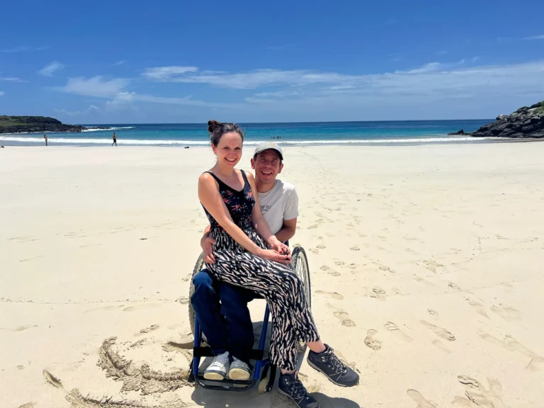 A woman sits on a mans lap in a wheelchair on a sandy beach, both smiling at the camera. The sky is blue with scattered clouds, and the ocean is calm in the background. Footprints lead across the sand.