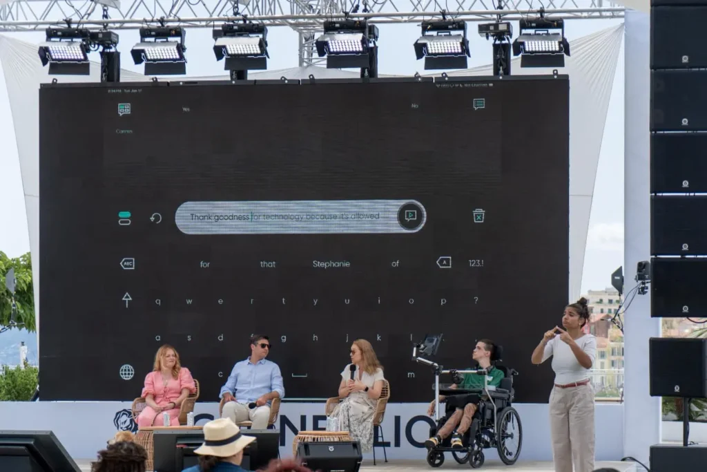 A large outdoor conference or festival stage setup with professional lighting rigs. On stage, there's a panel discussion taking place with several people seated in chairs, including one person in a wheelchair. There's a large screen displaying what appears to be a communication interface or app. A person is standing at the right side of the stage, likely presenting or moderating. The setting appears to be at a major conference or festival venue.