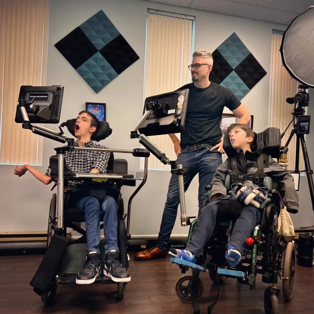 Three people in what appears to be a therapy or clinical setting with professional lighting equipment, showing three people from a wider angle. The geometric wall art is more prominently displayed, and there are adjustable tablet or communication device stands positioned in front of each wheelchair user. Professional video lighting equipment is visible.