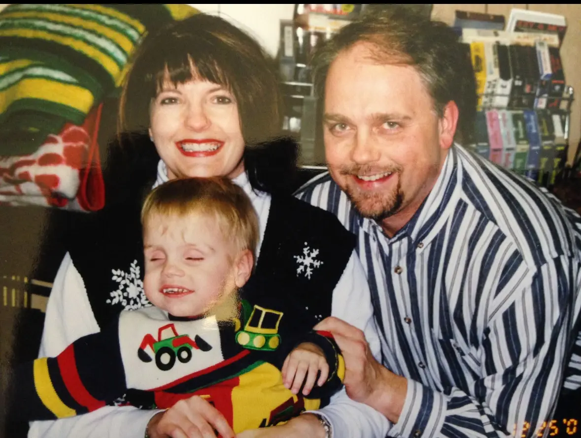 A family photo showing a woman with dark hair and bangs wearing a black top with snowflake decorations, a young blonde child in a colorful shirt with truck designs, and a man in a striped shirt. This appears to be an older photo, possibly from the late 1990s or early 2000s based on the image quality and styling.