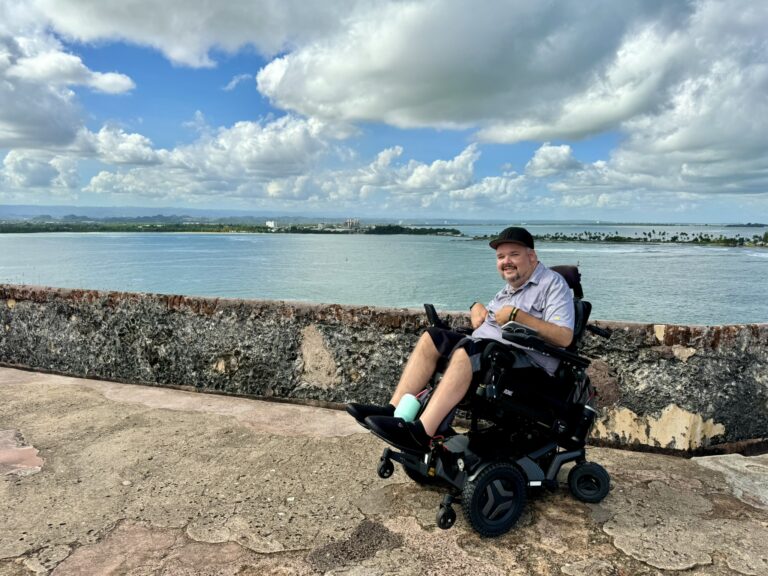 A smiling man in a power wheelchair sits on a stone walkway overlooking calm water and a distant shoreline under a blue sky with puffy clouds.