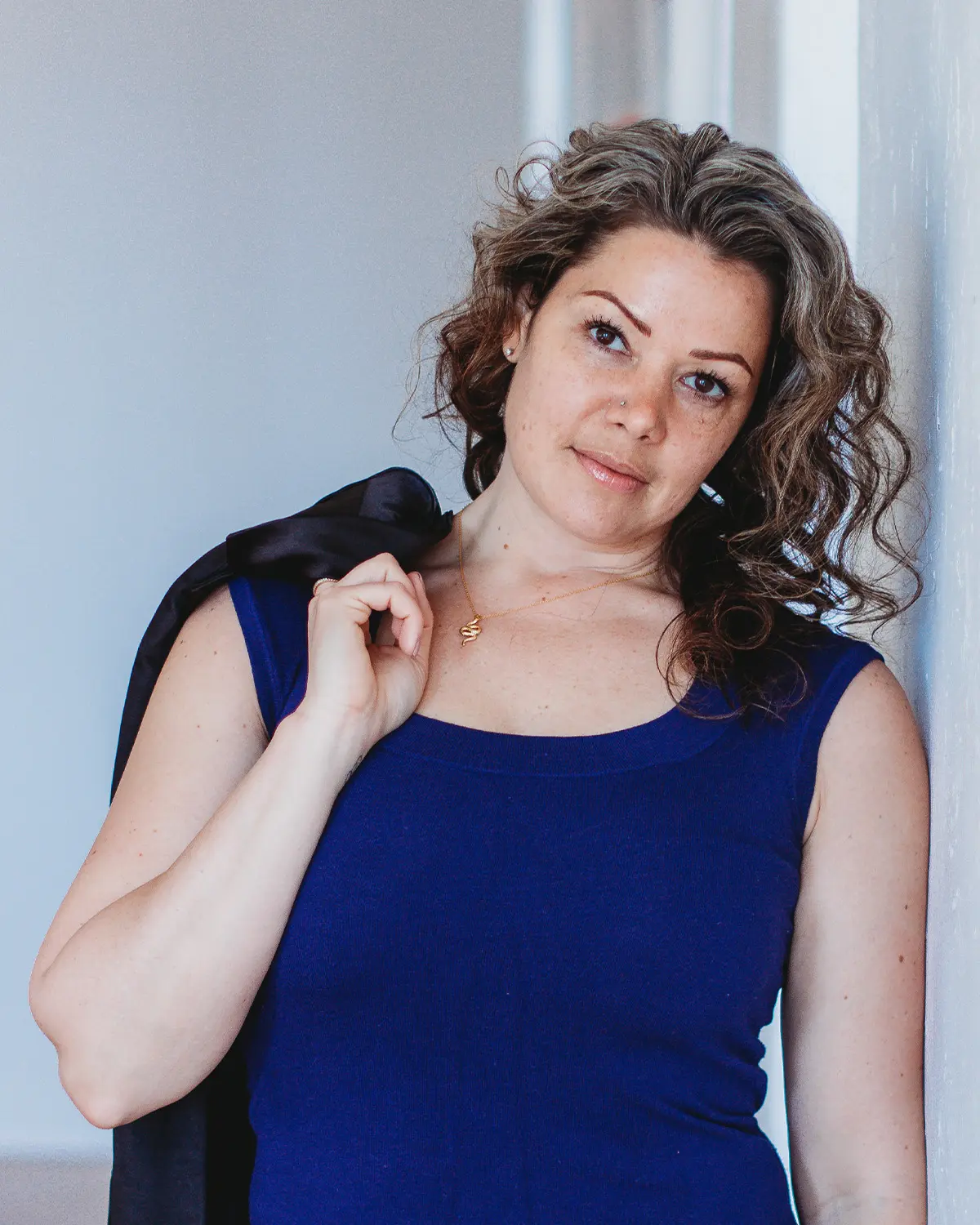 A woman with curly hair poses against a light wall, wearing a navy blue sleeveless top and holding a black jacket over her shoulder.