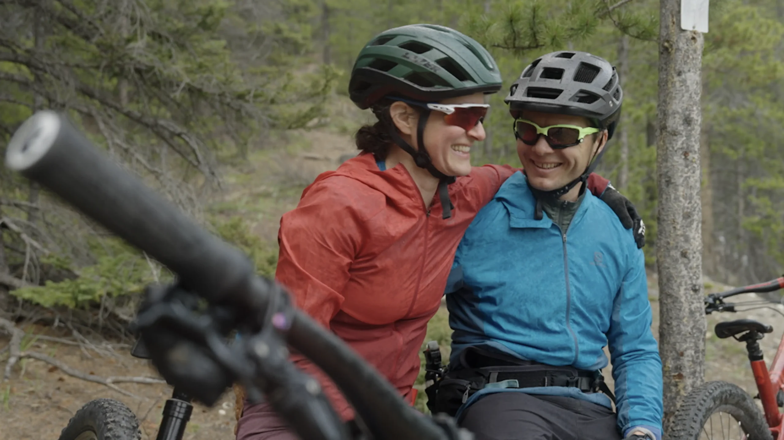 Two smiling people wearing cycling helmets and outdoor gear pose together on a forest trail. The person on the left wears a red jacket and green helmet, while the person on the right wears a blue jacket and gray helmet. A mountain bike is visible in the foreground, suggesting they're on a mountain biking adventure.