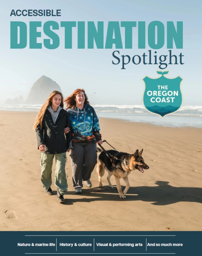 Two women, one with a guide dog, walk along a sandy beach on the Oregon Coast with Haystack Rock in the background, promoting accessible travel destinations.
