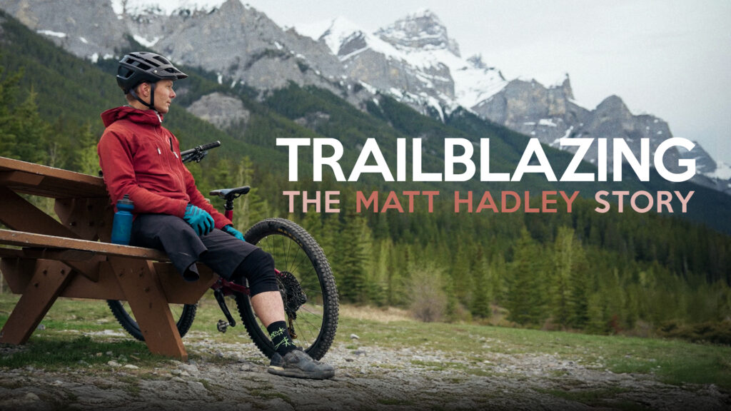 A mountain biker relaxes on a wooden bench, surrounded by lush greenery and majestic mountains, with a bicycle nearby.
