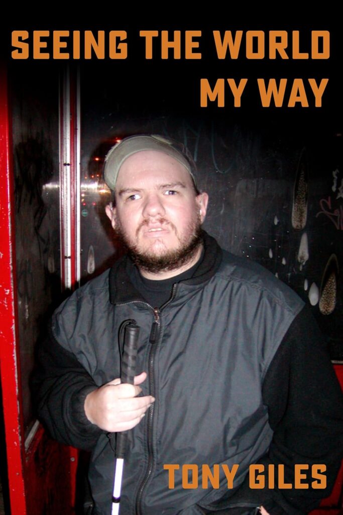 The cover of "Seeing The World My Way" by Tony Giles. A close-up photo of a man with a beard and a baseball cap holding a white cane. He is wearing a dark jacket and standing in front of a dark, graffiti-covered wall.