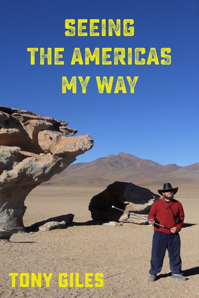 The cover of "Seeing The Americas My Way" by Tony Giles. A man with a beard, a cowboy hat, and a white cane stands in a barren desert landscape. A large, sculpted rock formation is to his left, and mountains are in the distance.