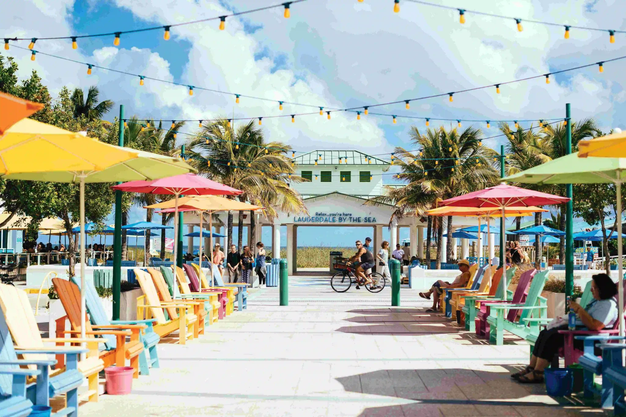 The image shows a vibrant, sunny plaza lined with colorful Adirondack chairs under equally colorful umbrellas. Palm trees frame the scene, and strings of yellow lights are strung overhead. In the center background, there's a white structure with a green roof that reads "Relax… you’re here LAUDERDALE BY THE SEA," opening up to a view of the ocean. People are strolling, chatting, and biking, giving the area a relaxed beach-town vibe.