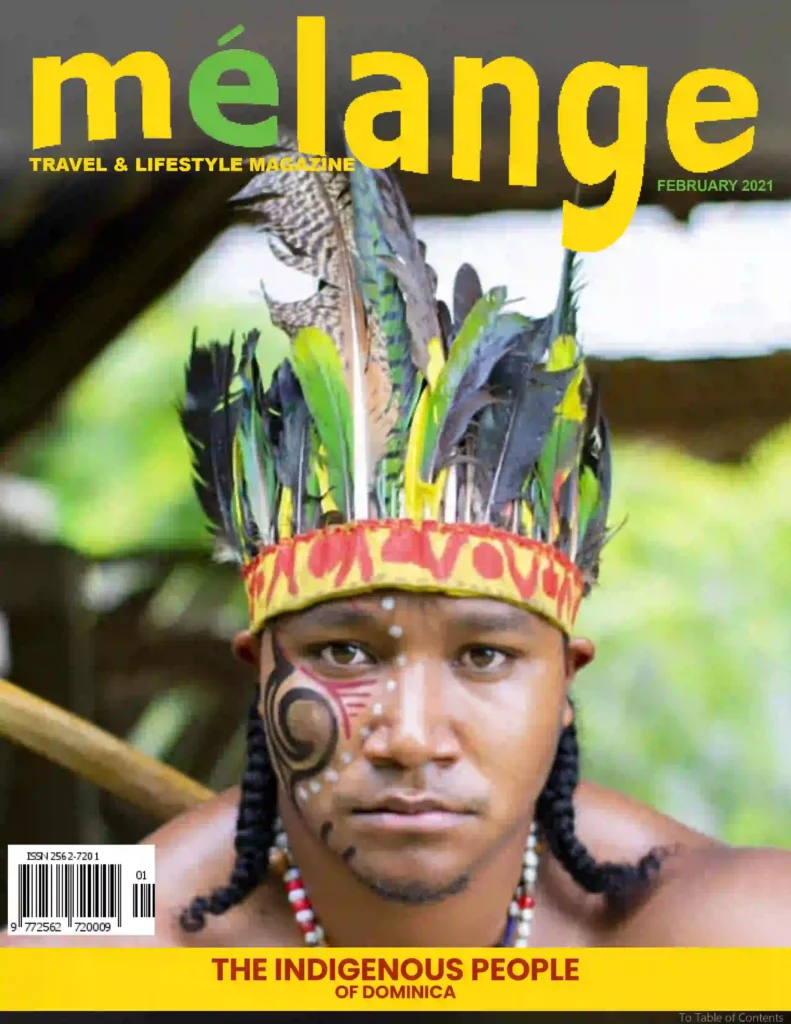 A close-up portrait of an Indigenous Kalinago man from Dominica, wearing a colorful feathered headdress and traditional face paint, featured on the cover of Mélange Travel & Lifestyle Magazine, February 2021 issue