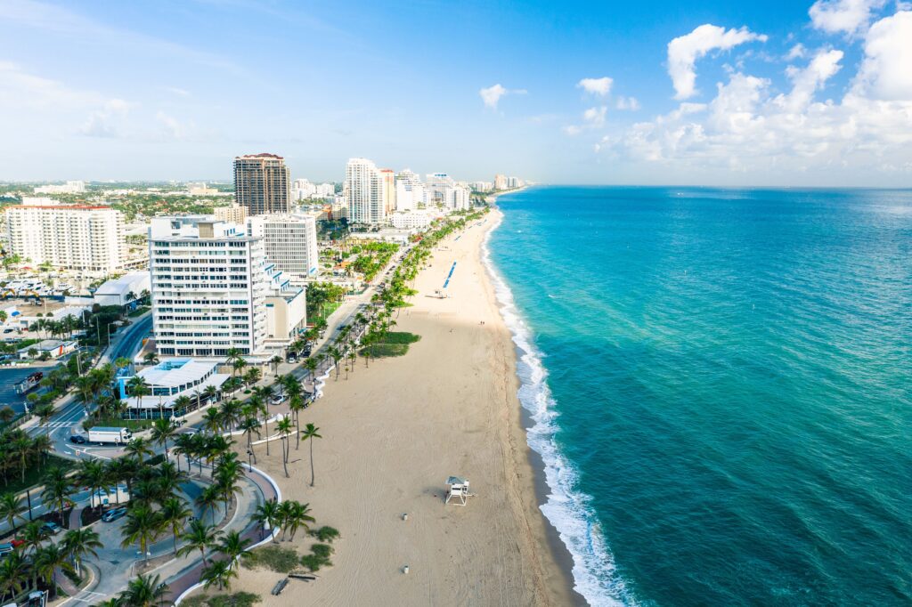 Aerial view of a sunny beach with turquoise water on the right and a city skyline with tall buildings and palm trees lining the shore on the left.