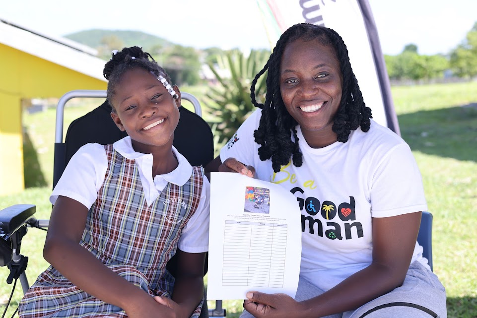 A smiling Black woman in a white "Be a Good Human" t-shirt sits outdoors next to a young girl in a school uniform and a motorized wheelchair. They are both looking at the camera and holding a sheet of paper with a chart and an image featuring a wheelchair symbol.