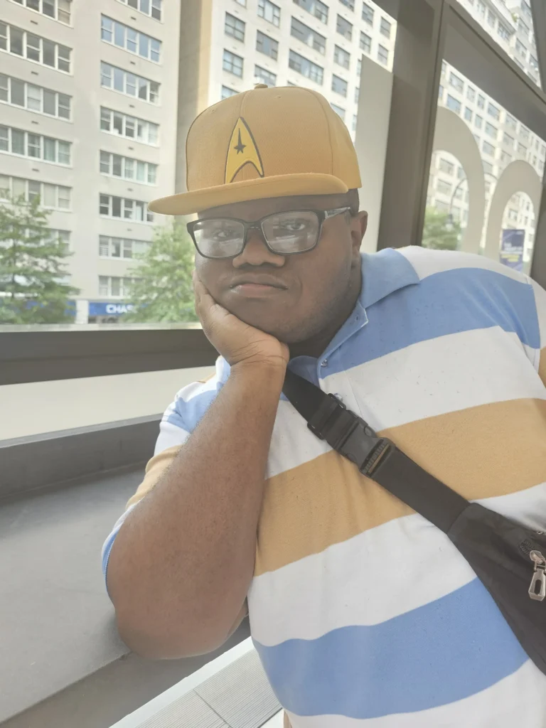 Man wearing a yellow Star Trek baseball cap, glasses, and a striped polo shirt, resting his chin on his hand with a city building visible through a large window behind him.