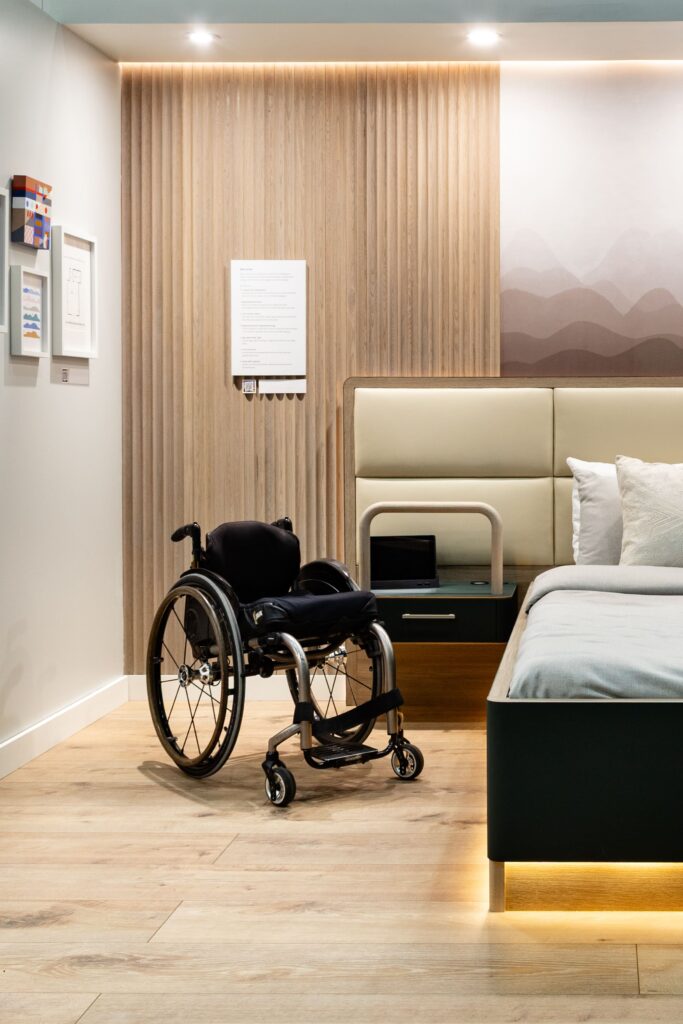 A modern, black wheelchair is parked in the center of an accessible bedroom suite. The room features light wood flooring and a wall with vertically slatted wood paneling behind a bed with a beige, rectangular upholstered headboard. A light is visible beneath the bed, and a laptop rests on a bedside table. On the wall to the left, a framed collection of small, colorful art pieces hangs near a vertical informational sign.