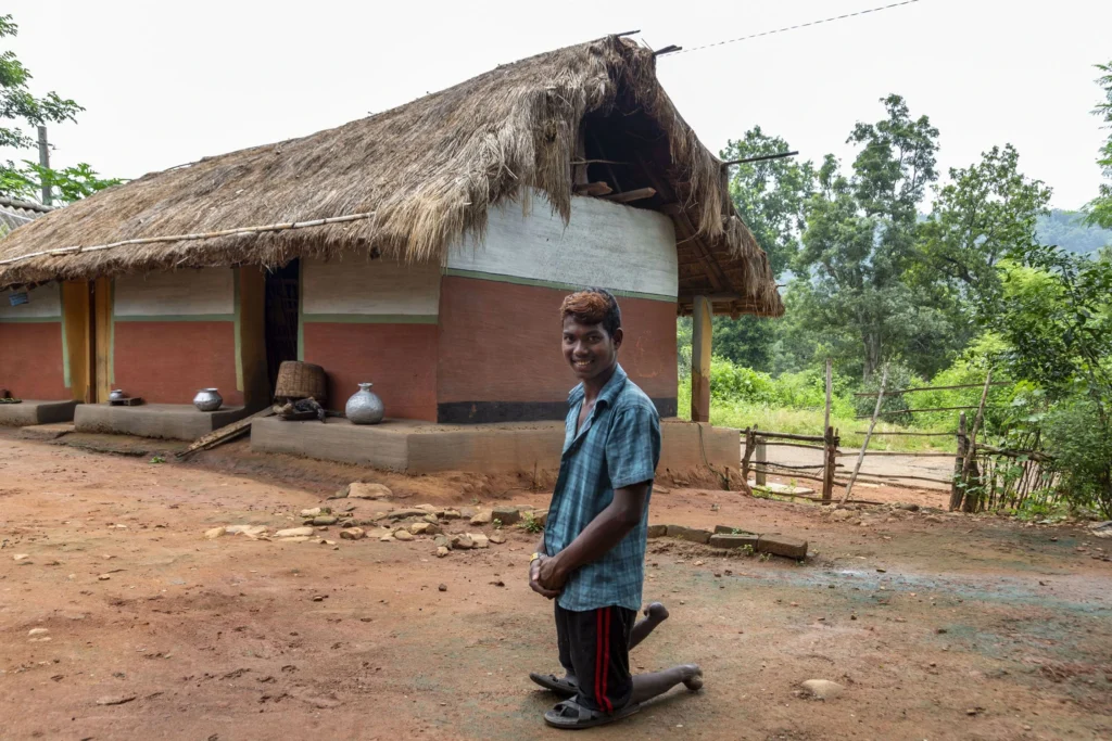 A young man named Chhotray Hembram stands on his knees, smiling at the camera, in front of a traditional rural home with a thatched roof and earthen walls. He is wearing a plaid blue and white shirt and black and red shorts. The house is set in an open area with dirt ground and lush greenery in the distance.