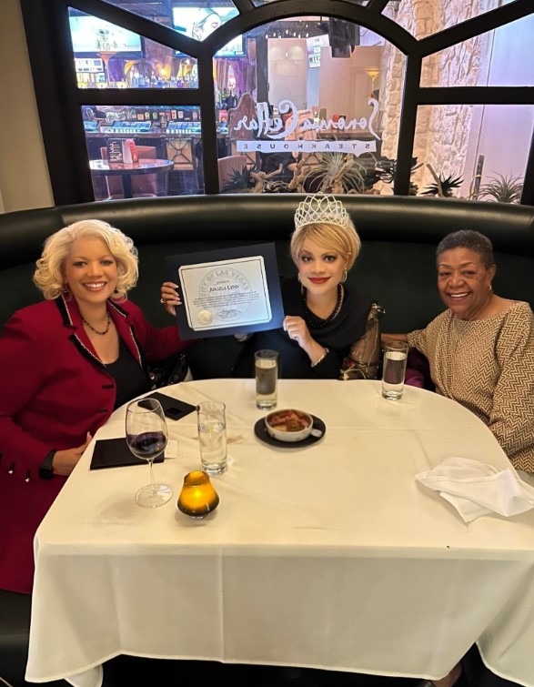 Angela Lynn, wearing a crown, proudly holds a certificate of honor from the City of Las Vegas while seated at a restaurant table with two women. The group smiles together in celebration of her recognition as the first deaf woman to win a state title in Ms. Corporate America.