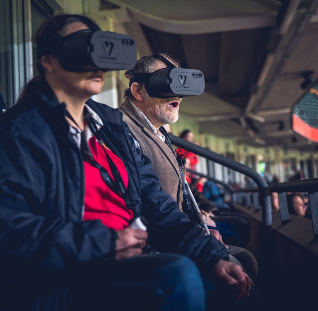 Two spectators, a woman and an older man, are seated side-by-side in a stadium wearing GiveVision smart headsets (or similar visual aids) over their eyes. The man appears to be reacting enthusiastically to the event, while the woman looks focused. The scene is dimly lit inside the stadium seating area, with a railing in the foreground.