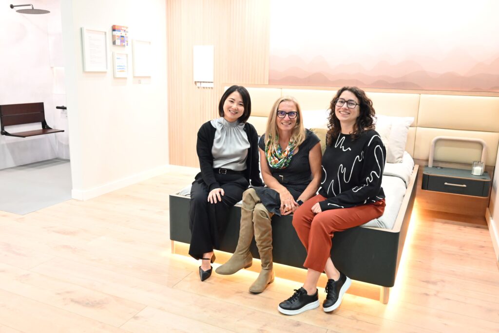 Three women, identified as Hannah, Julie, and Kaly, are sitting casually on the edge of the low, dark-framed bed in the accessible suite exhibit. They are posing and smiling at the camera. The setting is modern and brightly lit, featuring light wood floors and a cream-colored upholstered headboard against a wall with a subtle, wavelike gradient pattern.