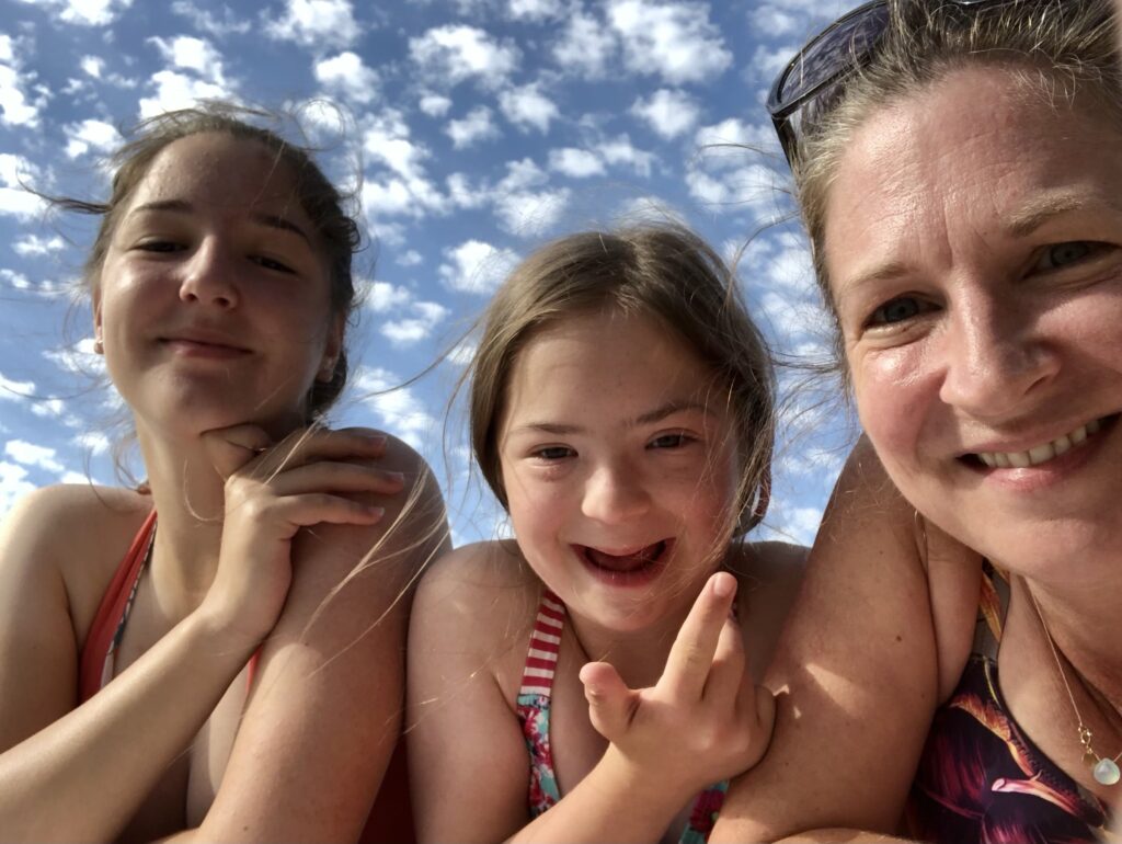 A sunny, close-up selfie of three females (likely a mother and two daughters) outside against a bright blue sky with wispy white clouds. The person on the right is smiling at the camera, and the young girl in the middle is also smiling widely, holding up her fingers.