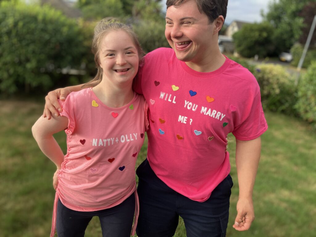 A bright, outdoor photo of a young man and woman with Down syndrome, standing side-by-side in a grassy yard. The man is wearing a bright pink t-shirt that says "WILL YOU MARRY ME?" and the woman is wearing a lighter pink t-shirt that says "NATTY & OLLY," both decorated with multicolored heart stickers. They are both smiling and looking at the camera.