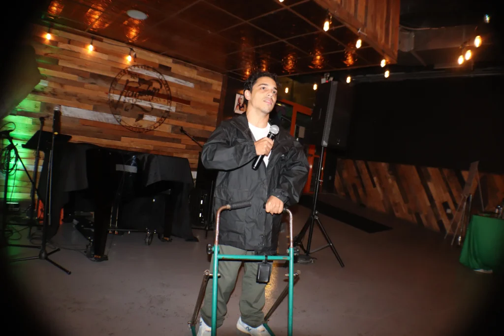 A young man is standing on a stage, holding a microphone and speaking into it while leaning on a green metal walker. He is wearing a black rain jacket and olive green pants. The stage has a rustic wood-paneled wall backdrop with an indistinct logo, and the ceiling has string lights. Music equipment and speakers are visible around the stage.