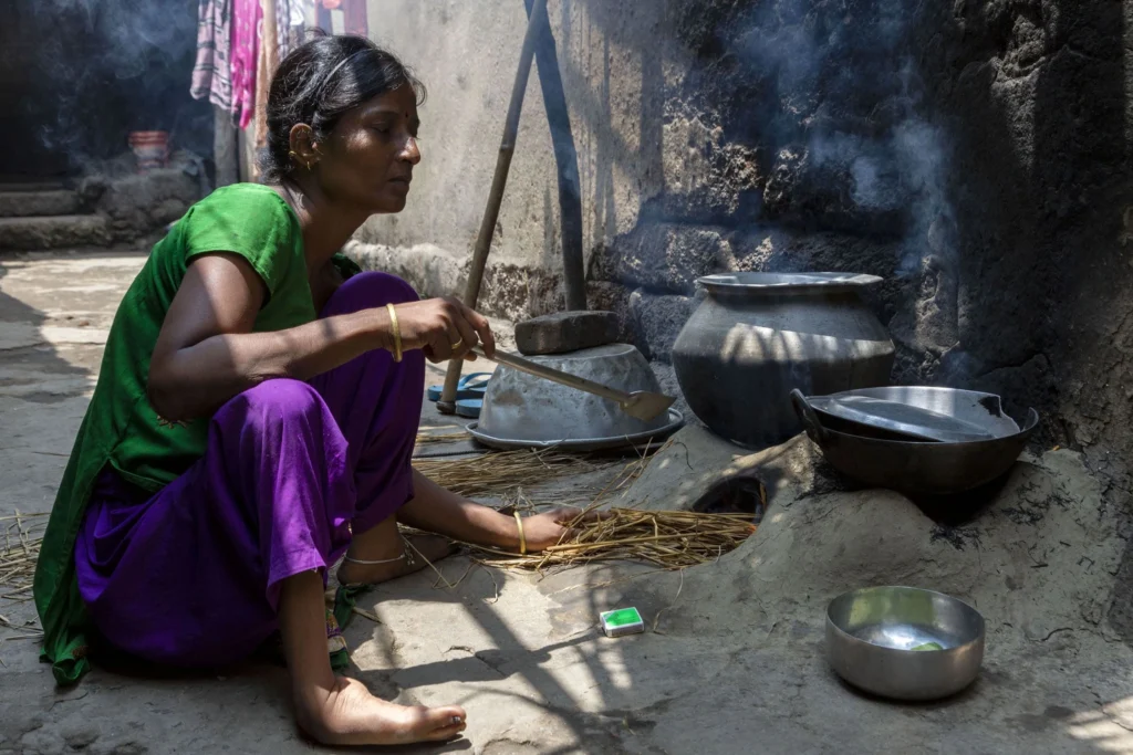 A woman named Koushalya Swain wearing a bright green top and vibrant purple pants crouches low, cooking outdoors over a traditional earthen stove (chulha). She is adding straw or kindling to the fire and holding a wooden spatula. Smoke rises from the cooking area. Nearby, a large earthen pot and a metal bowl sit on the ground.