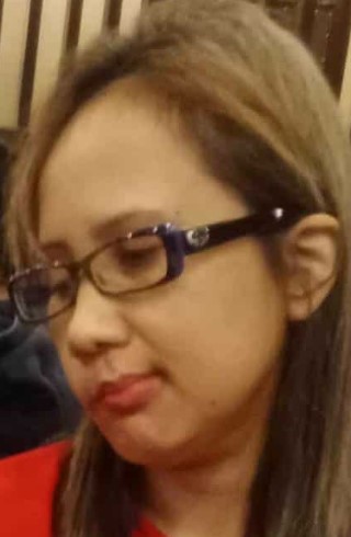 A close-up headshot of a woman looking down and to her right. She has light brown hair with blonde highlights, is wearing rectangular dark-rimmed eyeglasses with a purple accent on the temple, and a red shirt.