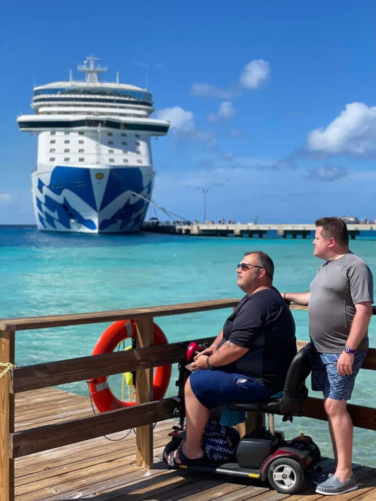 Two men on a wooden pier overlooking bright turquoise water. One man is seated in a dark red and black motorized scooter, wearing sunglasses and a navy shirt and shorts. The other man is standing beside him, wearing a gray t-shirt and blue patterned shorts. In the background, a large cruise ship with a distinctive blue and white patterned hull is docked at a longer pier under a sunny, blue sky.
