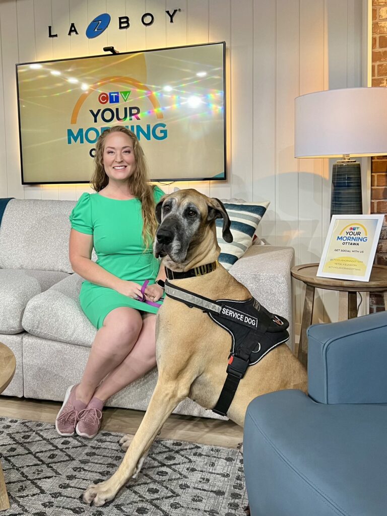 A photo of Michelle Weger and her service dog, Quinn, on the set of CTV's Your Morning show. Michelle is seated on a couch, smiling and wearing a bright green dress and pink running shoes. Quinn, the large Great Dane service dog wearing a black harness, is seated politely next to her. A monitor in the background displays the show's logo, and the set features comfortable furniture and a patterned rug.
